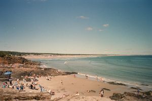 Ogunquit Beach in Maine seen from the rocks along Marginal Way, sunbathers and umbrellas scattered across the sand, surf breaking into a wide blue bay under a clear summer sky